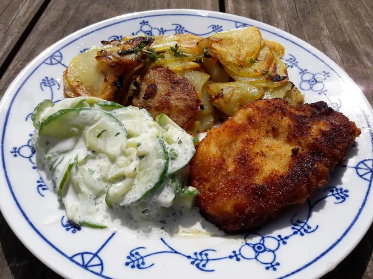 Ein knuspriges Schnitzel mit goldbraunen Ofenkartoffeln und einem cremigen Gurkensalat auf einem blauen Zwiebelmuster-Teller.