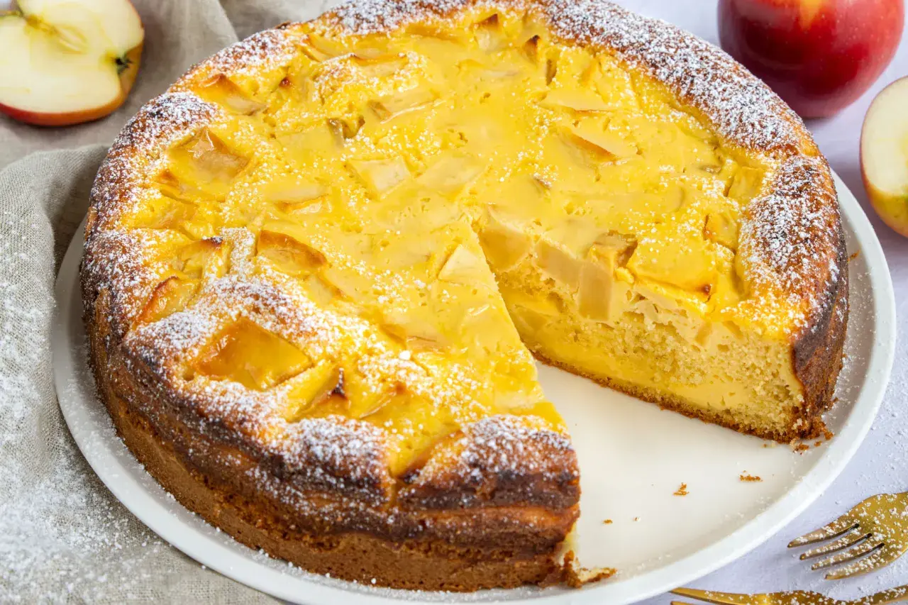Ein schneller, saftiger Apfelkuchen mit Quark, bestäubt mit Puderzucker. Daneben liegen Äpfel und goldene Gabeln.