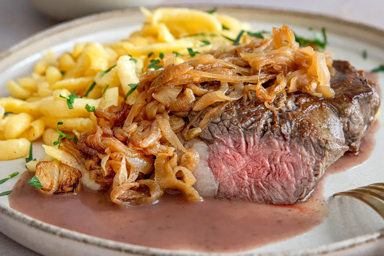 Zwiebelrostbraten im Backofen, saftig rosa gebraten, mit goldbraunen Röstzwiebeln und Spätzle auf einem Teller.