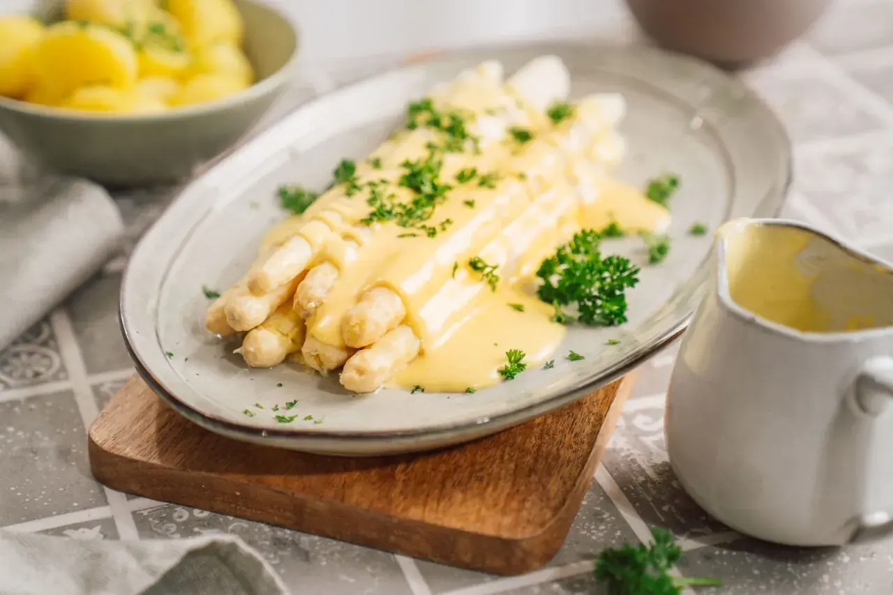 Spargel mit cremiger Sauce Hollandaise, ein Klassiker, der mit diesem Rezept gelingt. Beilagen wie Kartoffeln sind im Hintergrund zu sehen.
