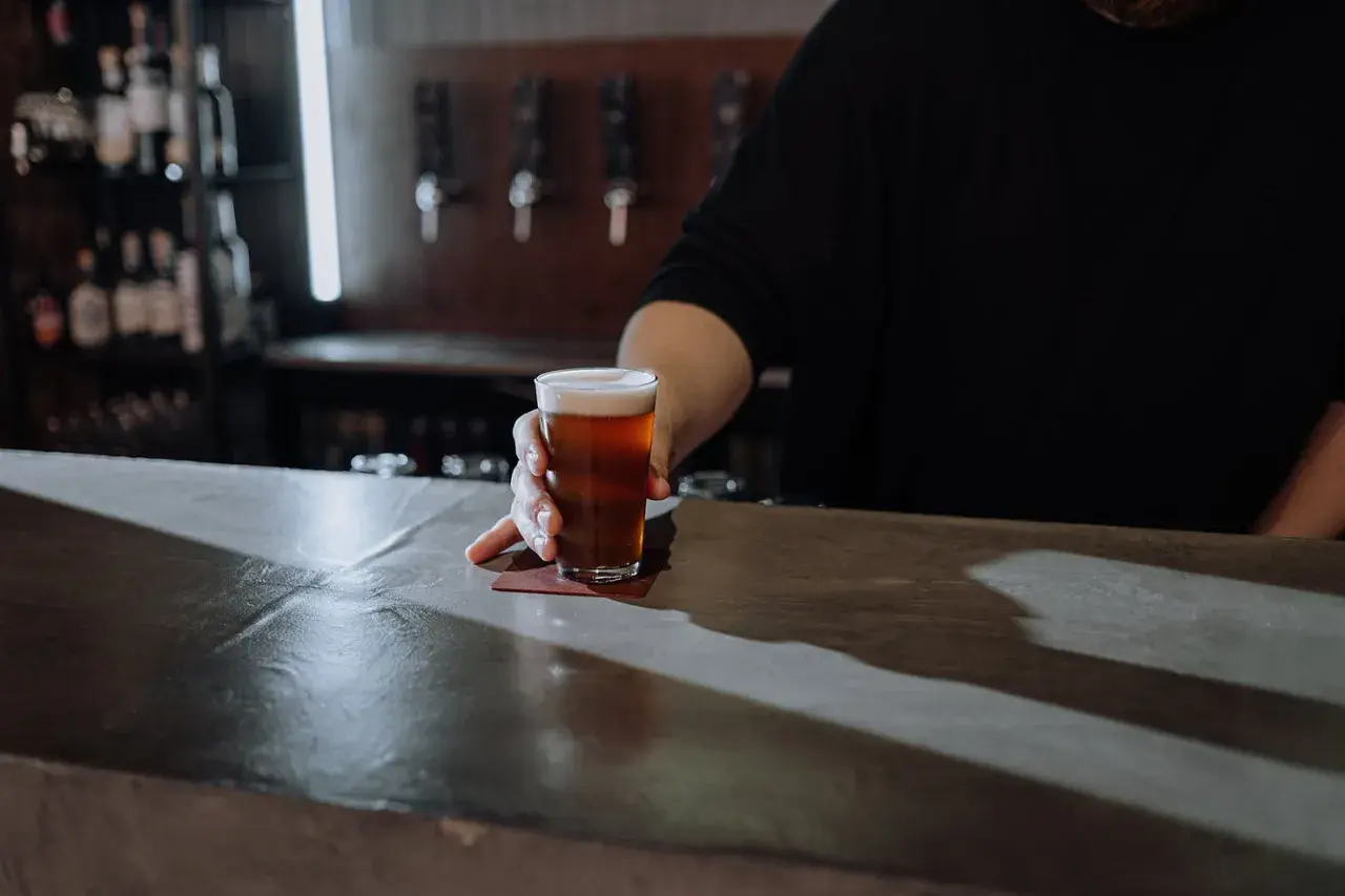 Ein Glas Bier wird über den Tresen geschoben. Der Unterschied zwischen Bier und Pils ist hier nicht ersichtlich, aber die Farbe des Bieres ist bernsteinfarben.