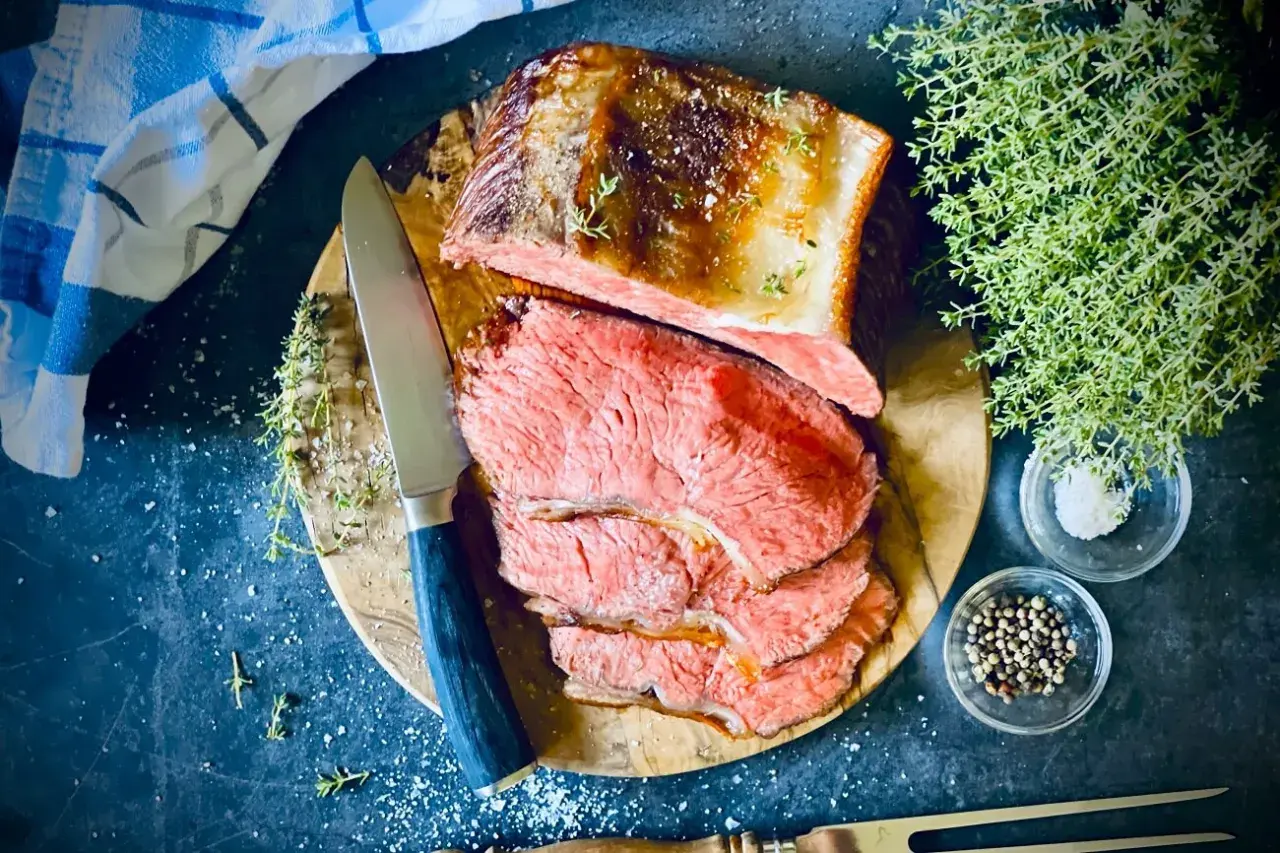 Saftiger Rostbraten, perfekt im Ofen zubereitet, liegt auf einem Holzbrett. Daneben liegen Kräuter, Salz und Pfefferkörner.
