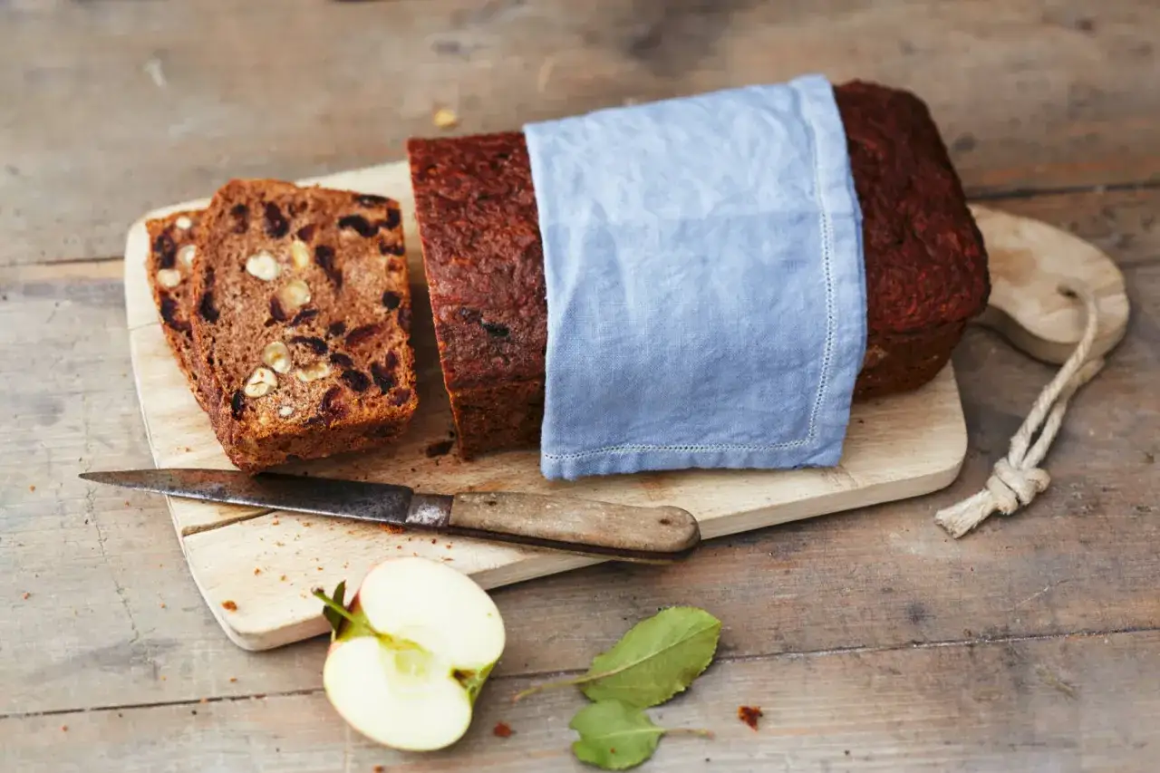 Ein saftiges Apfelbrot mit Nüssen und Rosinen, perfekt für das Apfelbrot Rezept. Daneben liegen zwei Scheiben und ein Messer.