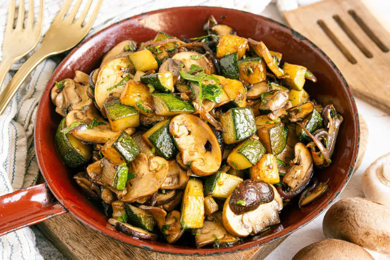 Herzhafte zucchini pilz pfanne, mit Kr&auml;utern verfeinert, dampft in einer roten Pfanne. Goldene Gabeln liegen bereit.