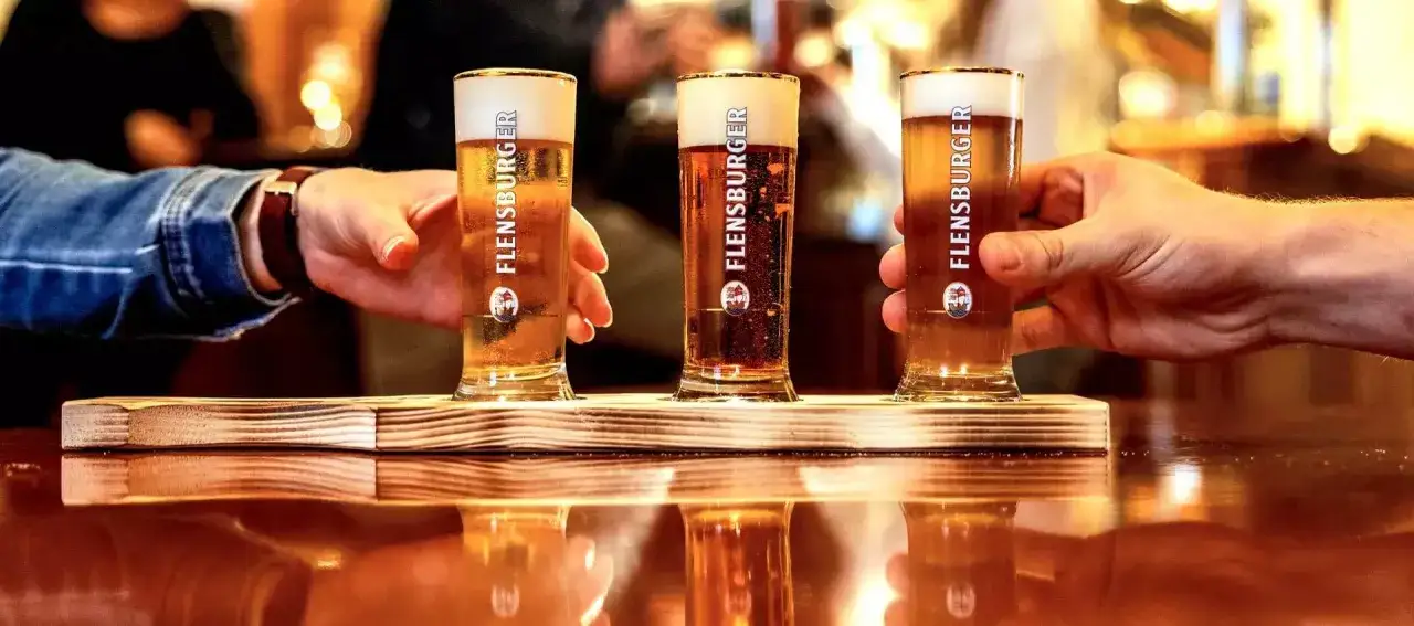 Drei Gl&auml;ser mit goldgelbem Bier und cremigem Bier schaum stehen auf einem Holzbrett. H&auml;nde greifen nach den Gl&auml;sern.