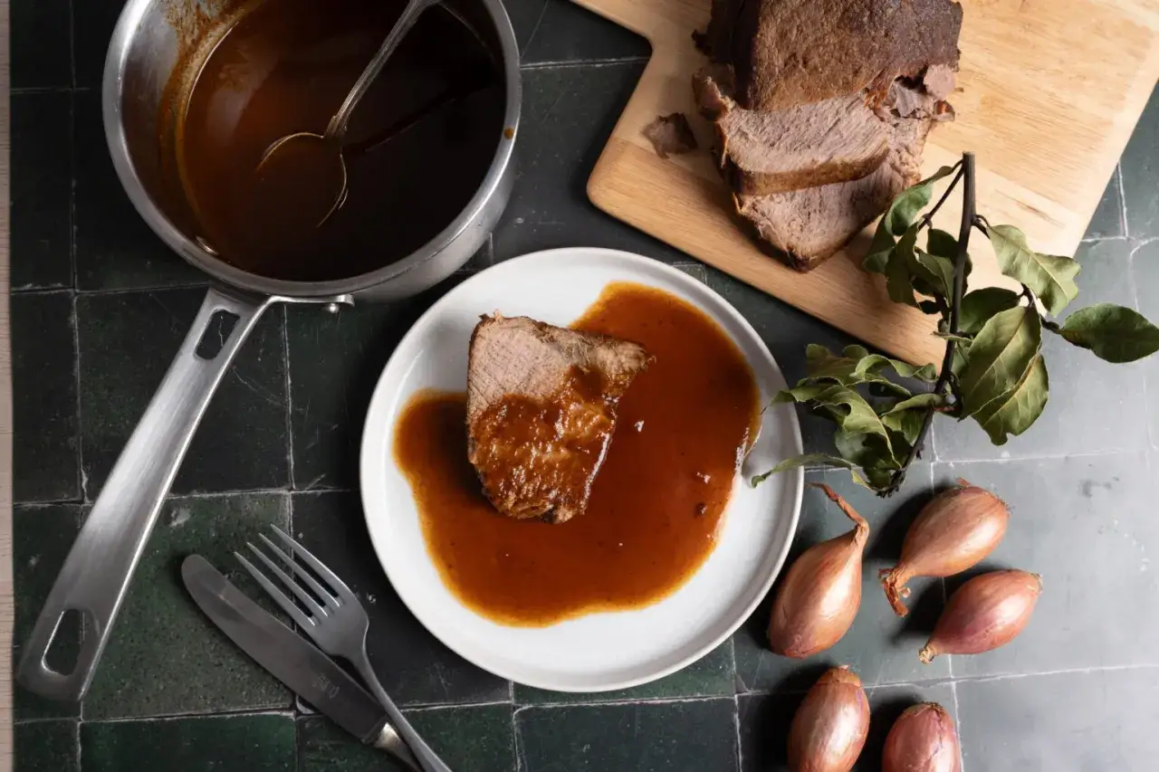 Ein St&uuml;ck Rinderbraten auf einem Teller, &uuml;bergossen mit einer k&ouml;stlichen Rinderbraten Sauce. Daneben ein Topf mit Sauce, Besteck und Schalotten.
