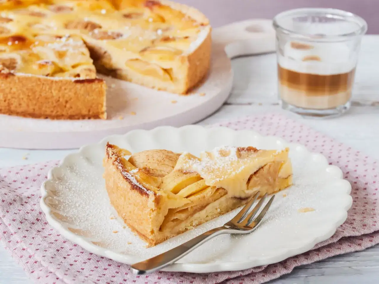 Ein St&uuml;ck saftiger Apfelkuchen mit Guss, best&auml;ubt mit Puderzucker, wartet auf dem Teller. Daneben ein Glas Latte Macchiato.