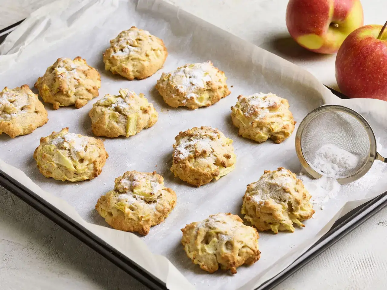 Saftige Apfelkekse, frisch gebacken und mit Puderzucker best&auml;ubt, liegen auf einem Backblech. Daneben zwei &Auml;pfel und ein Sieb mit Puderzucker.