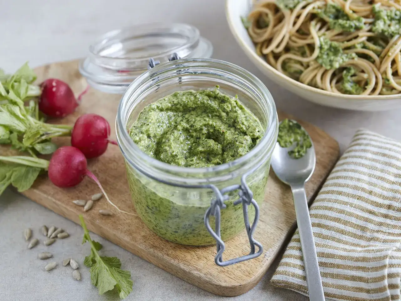 Frisches Radieschen Pesto im Glas, daneben Radieschen und Sonnenblumenkerne. Ein Teller Spaghetti mit Pesto ist im Hintergrund zu sehen.