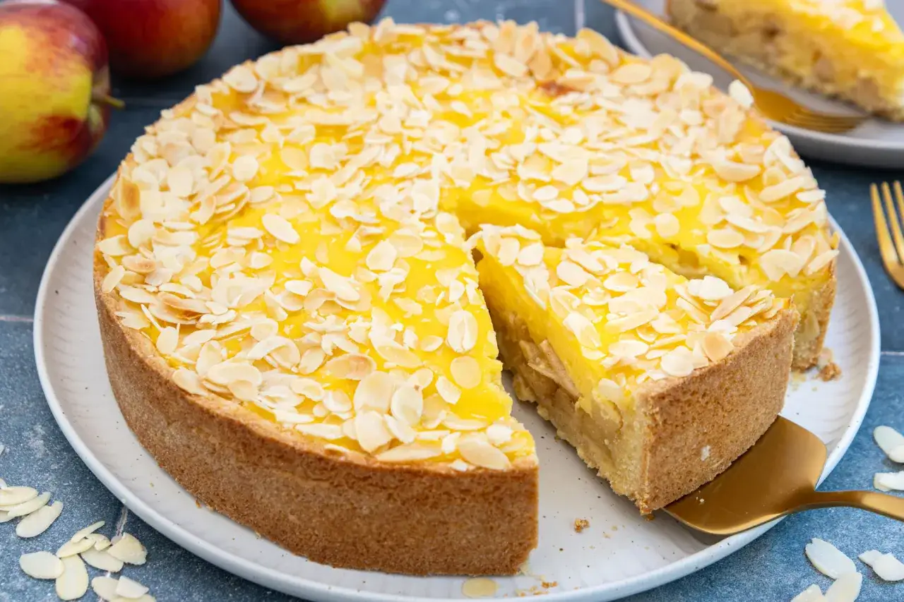 Ein k&ouml;stlicher versunkener Apfelkuchen mit Pudding und Mandelbl&auml;ttchen. Ein St&uuml;ck wird gerade mit einem goldenen Tortenheber herausgehoben.