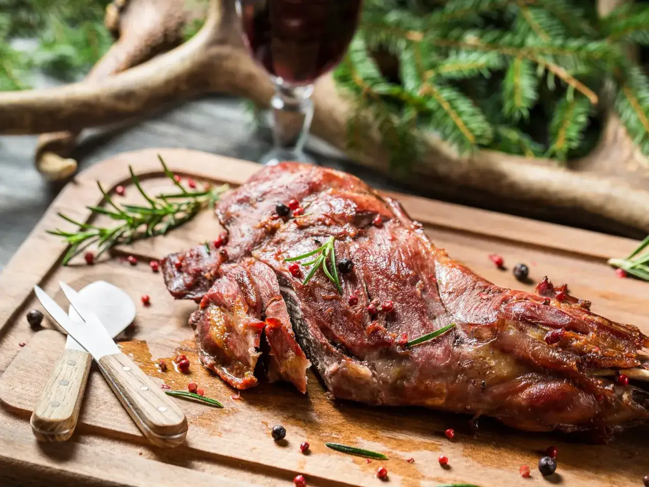 Ein saftiger Rehschlegel, perfekt gebraten, liegt auf einem Holzbrett. Gew&uuml;rze und Rosmarin zieren das Fleisch. Ein Glas Rotwein und Hirschgeweih im Hintergrund deuten auf ein k&ouml;stliches Rehschlegel Rezept hin.