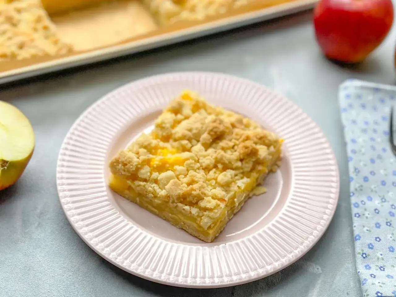 Ein Stück von Omas weltbester Apfelkuchen mit Streuseln auf einem rosa Teller, daneben ein halber Apfel und ein Serviertuch.