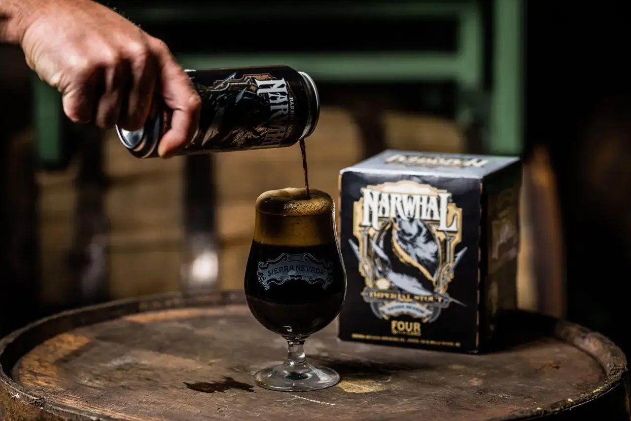 Ein Mann gie&szlig;t Sierra Nevada Narwhal Imperial Stout aus einer Dose in ein Glas. Das teure Bier steht neben einer Viererpackung auf einem alten Fass.
