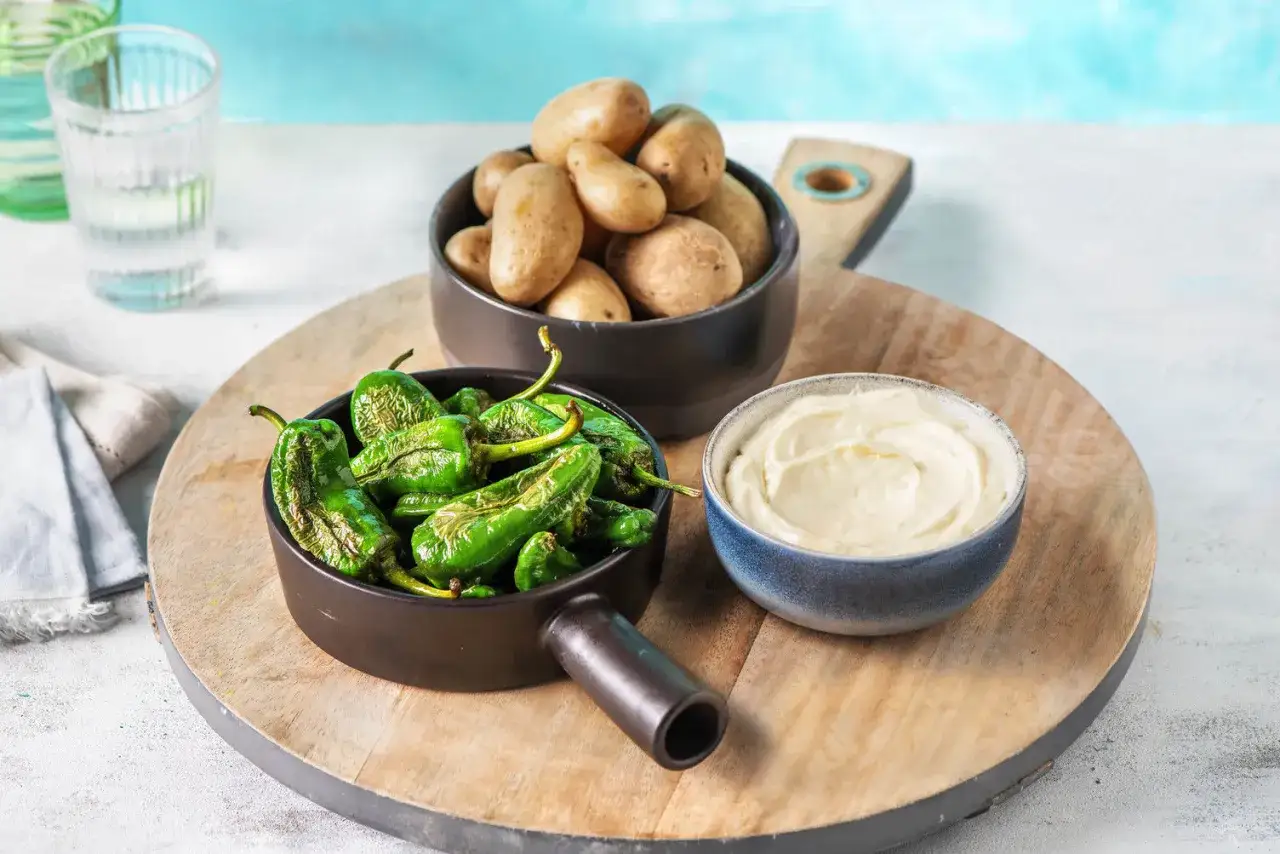 Pimientos de Padr&oacute;n rezept: Gebratene gr&uuml;ne Paprika, kleine Kartoffeln und eine cremige Dip-Sauce auf einem Holzbrett.