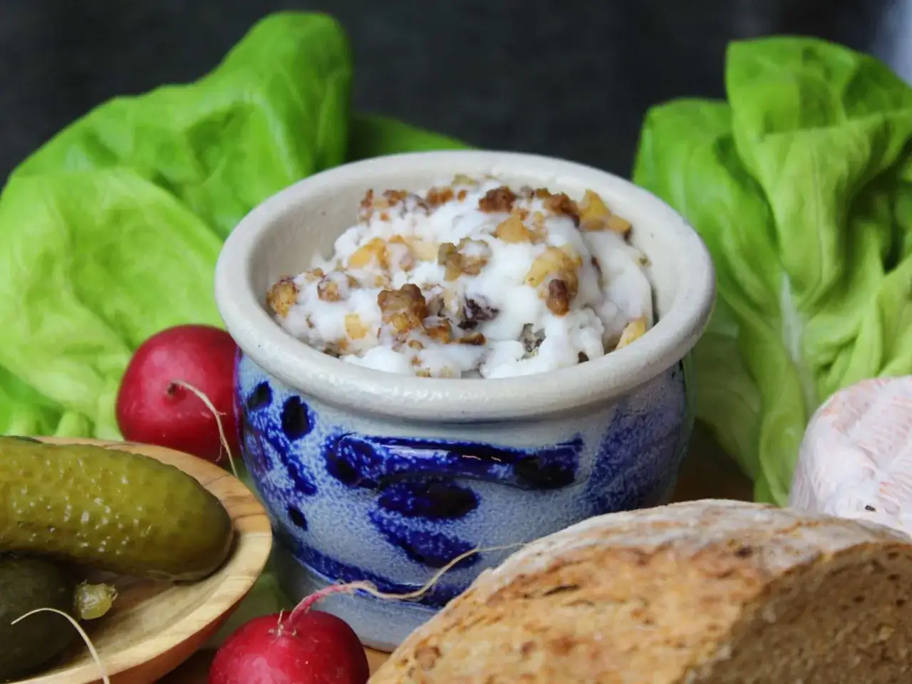 Schmalz mit knusprigen Grieben in einem blauen Steinguttopf, umgeben von Salat, Radieschen, Gew&uuml;rzgurken und Brot. Perfekt, um die Flomen auszulassen!