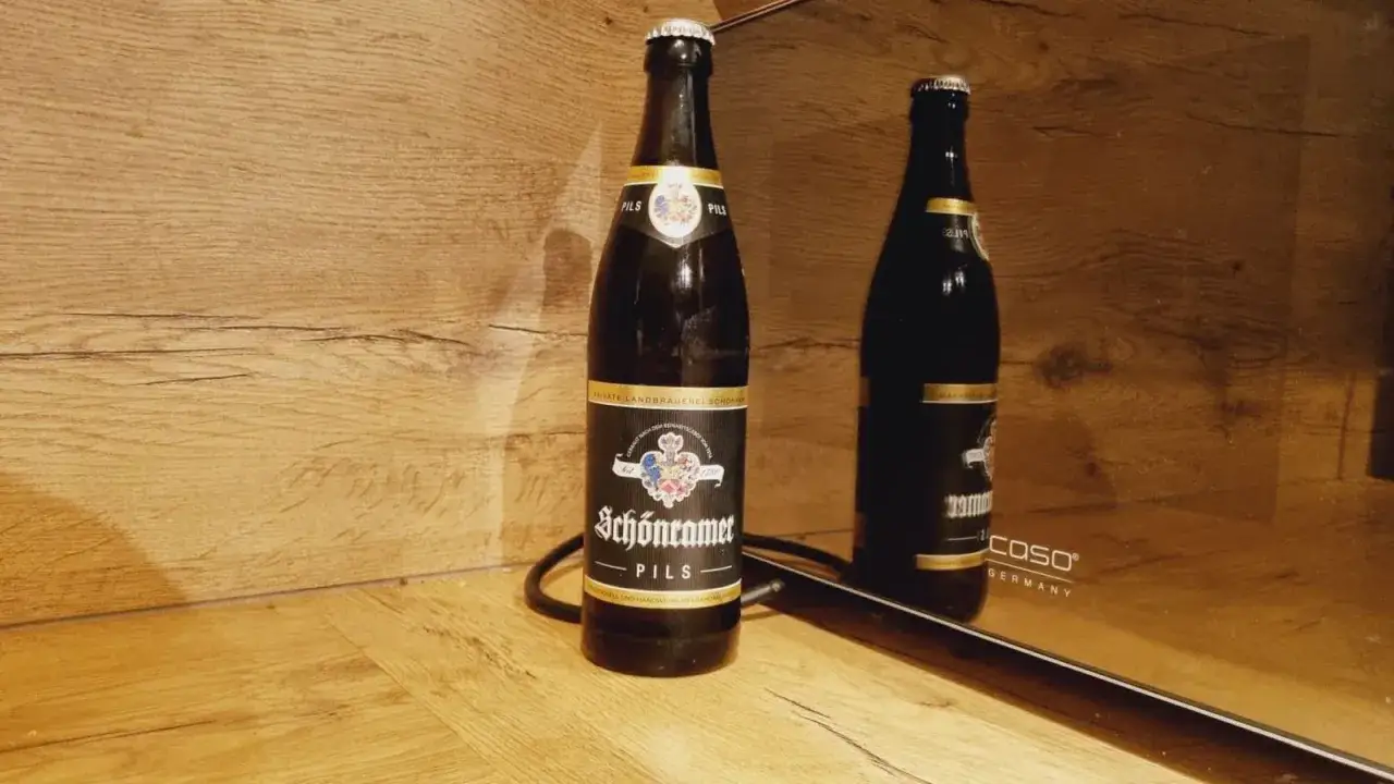 Sch&ouml;nramer Pils, ein Bier mit 35 IBU Hopfen, steht vor einer Holzvert&auml;felung. Sein Spiegelbild ist auf einer gl&auml;nzenden Oberfl&auml;che zu sehen.