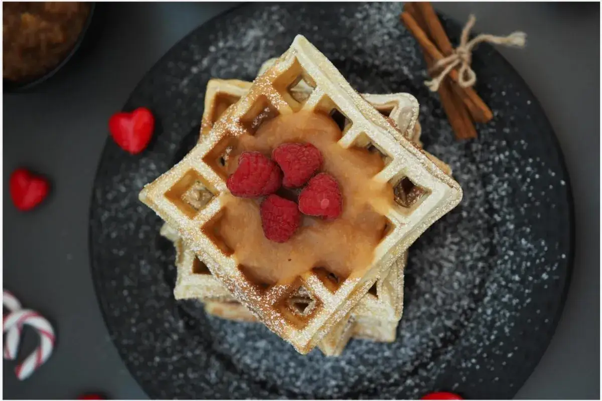 Gestapelte Waffeln mit Apfelmus und Himbeeren, best&auml;ubt mit Puderzucker. Daneben Zimtstangen und rote Herzen.