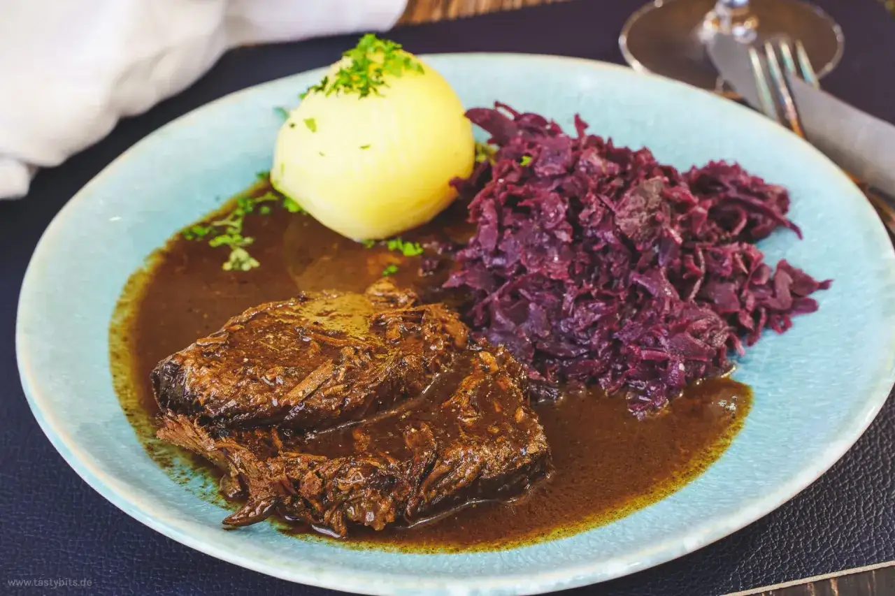 Zarter Rinderbraten in Rotwein eingelegt, serviert mit Kl&ouml;&szlig;en und Rotkohl auf einem blauen Teller.