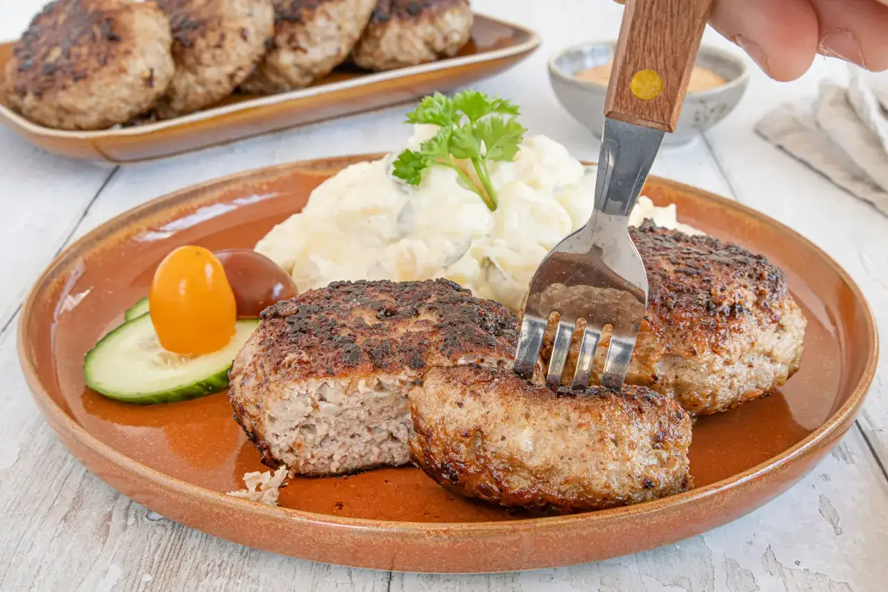 Beefsteak nach Omas Rezept: saftige Frikadellen mit Kartoffelsalat, Gurke und Tomate. Ein Gabel sticht in ein St&uuml;ck.