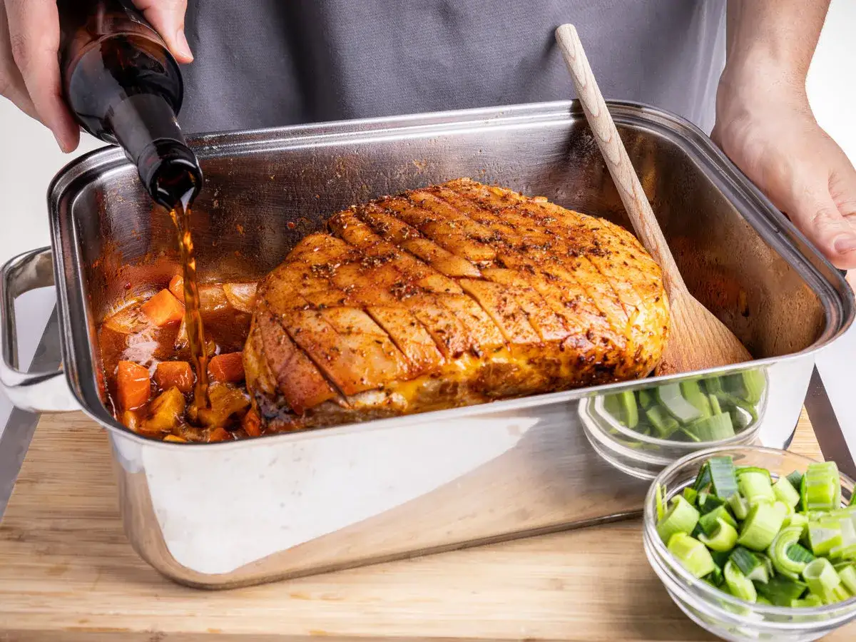 Ein Braten wird in einer Metallform mit Gem&uuml;se &uuml;bergossen. Die richtige schweinebraten temperatur umluft ist entscheidend f&uuml;r ein knuspriges Ergebnis.