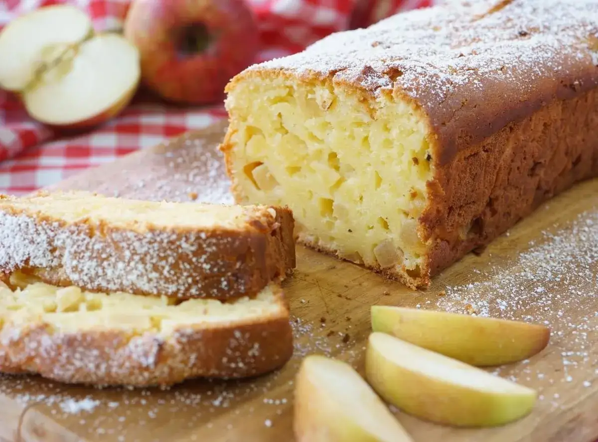 Ein schneller saftiger Apfelkuchen mit Quark, best&auml;ubt mit Puderzucker, neben frischen Apfelspalten und &Auml;pfeln auf kariertem Tuch.