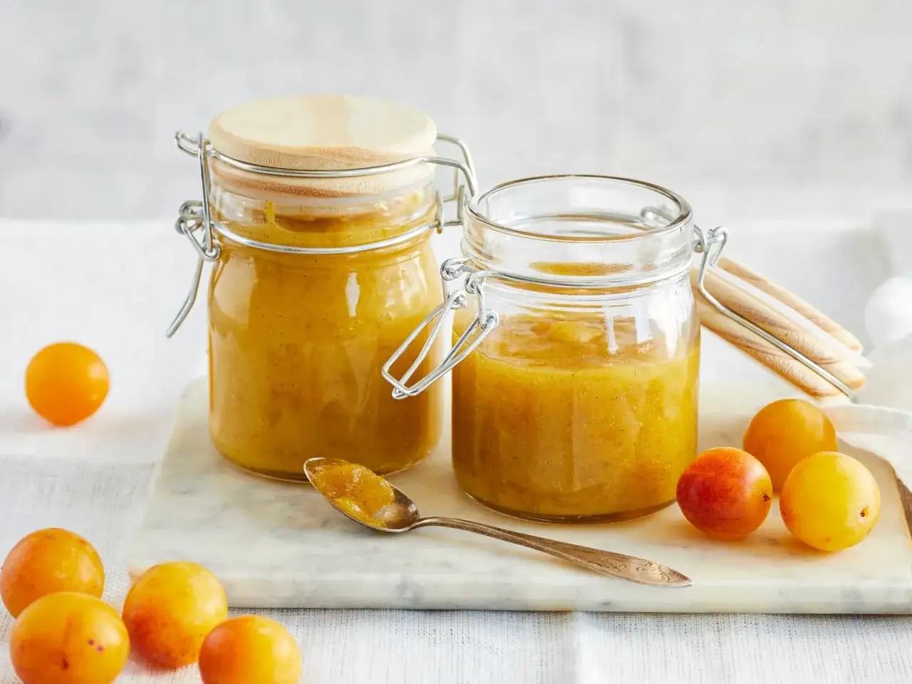 Zwei Gl&auml;ser mit goldgelber Mirabellenmarmelade, daneben frische Mirabellen und ein L&ouml;ffel voll Marmelade. Ein einfaches mirabellenmarmelade rezept.