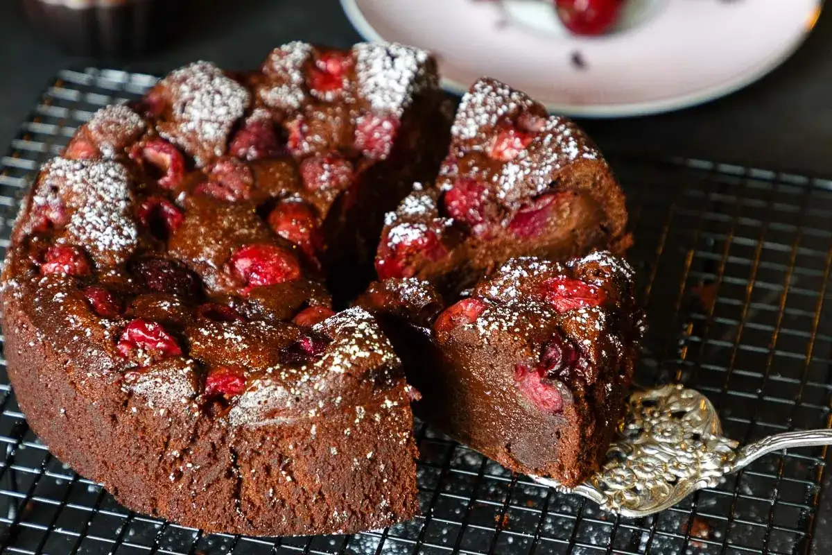 Saftiger Schoko-Kirsch-Kuchen aus der Springform, mit Puderzucker best&auml;ubt und Kirschen garniert. Ein St&uuml;ck wird gerade mit einer Tortengabel herausgehoben.