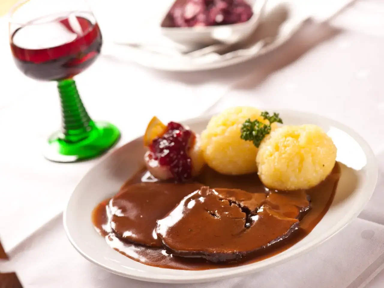 Rehbraten zubereiten im Backofen: Zarte Scheiben Rehfleisch in dunkler Sauce mit Kl&ouml;&szlig;en und Preiselbeeren, dazu ein Glas Rotwein.