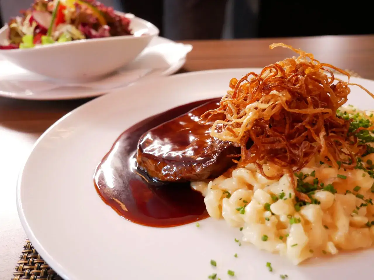 Zwiebelrostbraten im Backofen, serviert mit Spätzle und garniert mit knusprigen Röstzwiebeln und frischem Schnittlauch. Daneben ein bunter Salat.