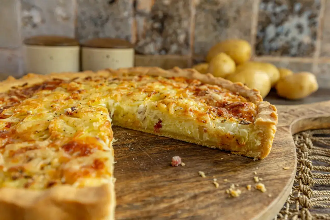 Ein goldbrauner Kartoffelkuchen vom Blech, mit K&auml;se &uuml;berbacken, liegt auf einem Holzbrett. Frische Kartoffeln im Hintergrund.