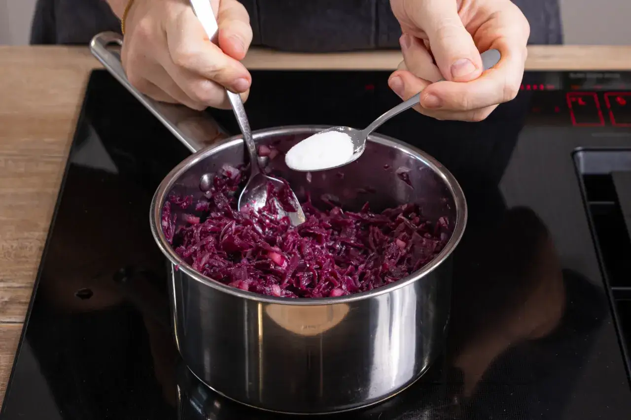 Zutaten f&uuml;r ein Rotkohl-Rezept aus dem Glas werden in einem Topf vermischt. Eine Hand gibt Zucker hinzu.