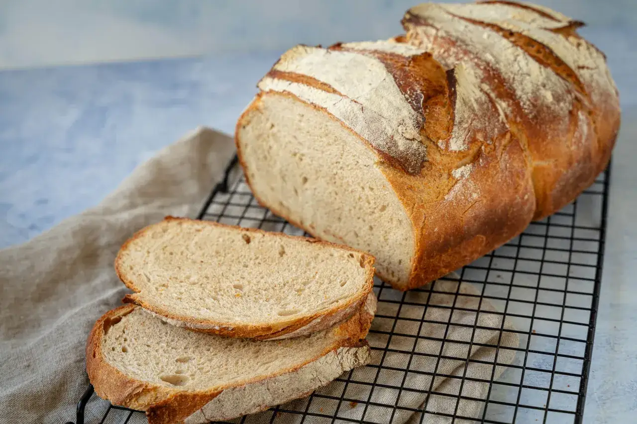 Ein frisch gebackenes Brot, das auf einem Gitterrost abkühlt. Zwei Scheiben liegen daneben. Perfekt für einfaches Brot backen.