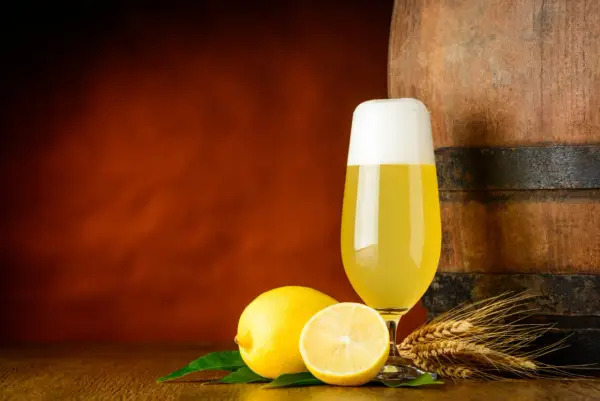 Ein Glas Radler steht neben Zitronen und Gerstenähren vor einem Holzfass.