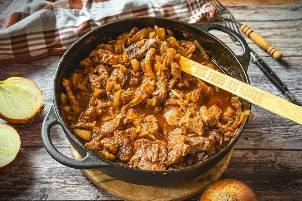 Saftiger Zwiebelbraten in einer Gusseisenpfanne mit Zwiebeln und Fleisch, daneben halbierte Zwiebeln.